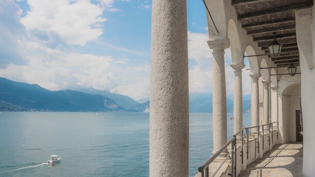 Ein Balkon am Wasser mit weißen Säulen und Steinboden, der auf einen stillen See mit einem Boot in der Ferne und Bergen im Hintergrund blickt.