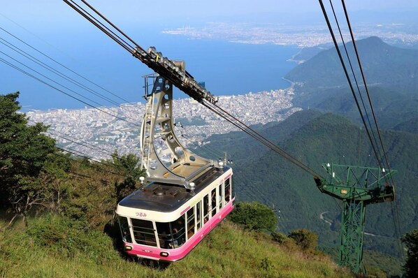 Un sistema de teleférico con un góndola rosa y blanca, un paisaje urbano al fondo y terreno montañoso.