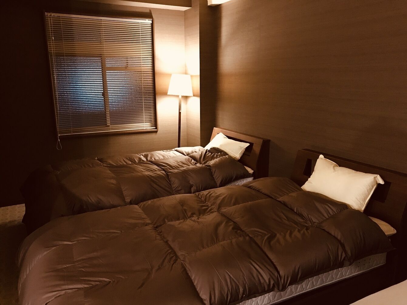 A hotel room with two beds, a bedside lamp, and a window with blinds.