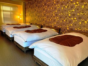 A hotel room with three beds, each with white linens and brown pillows. The room has a patterned wallpaper and a window with blinds. - Matsuki building Seaside Mansion (Beppu)