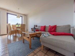 A cozy living area with a sofa, red cushions, a dining table with chairs, and a view through a large window.