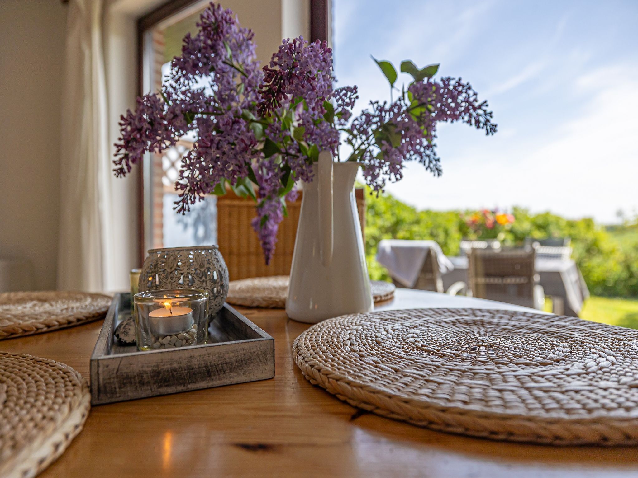Una mesa con un jarrón de lilas, una vela y una bandeja con vasos.
