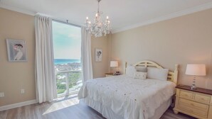 A bedroom with a large bed, a chandelier, a nightstand, and a view of the ocean. - BOOK YOUR BEACH VACATION! Penthouse Unit with Stunning Views of Destin! (Destin)