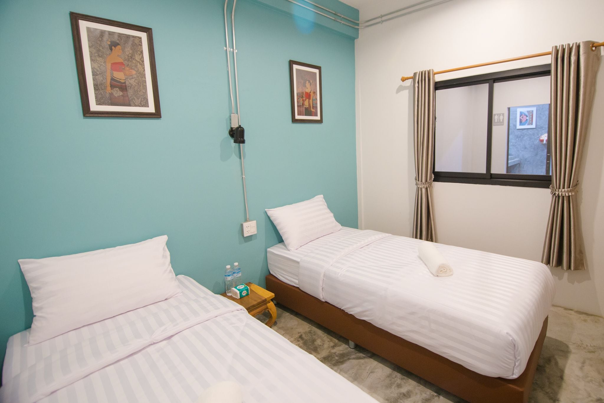 A hotel room with two single beds, a small wooden nightstand, a wall socket, and a window with curtains.