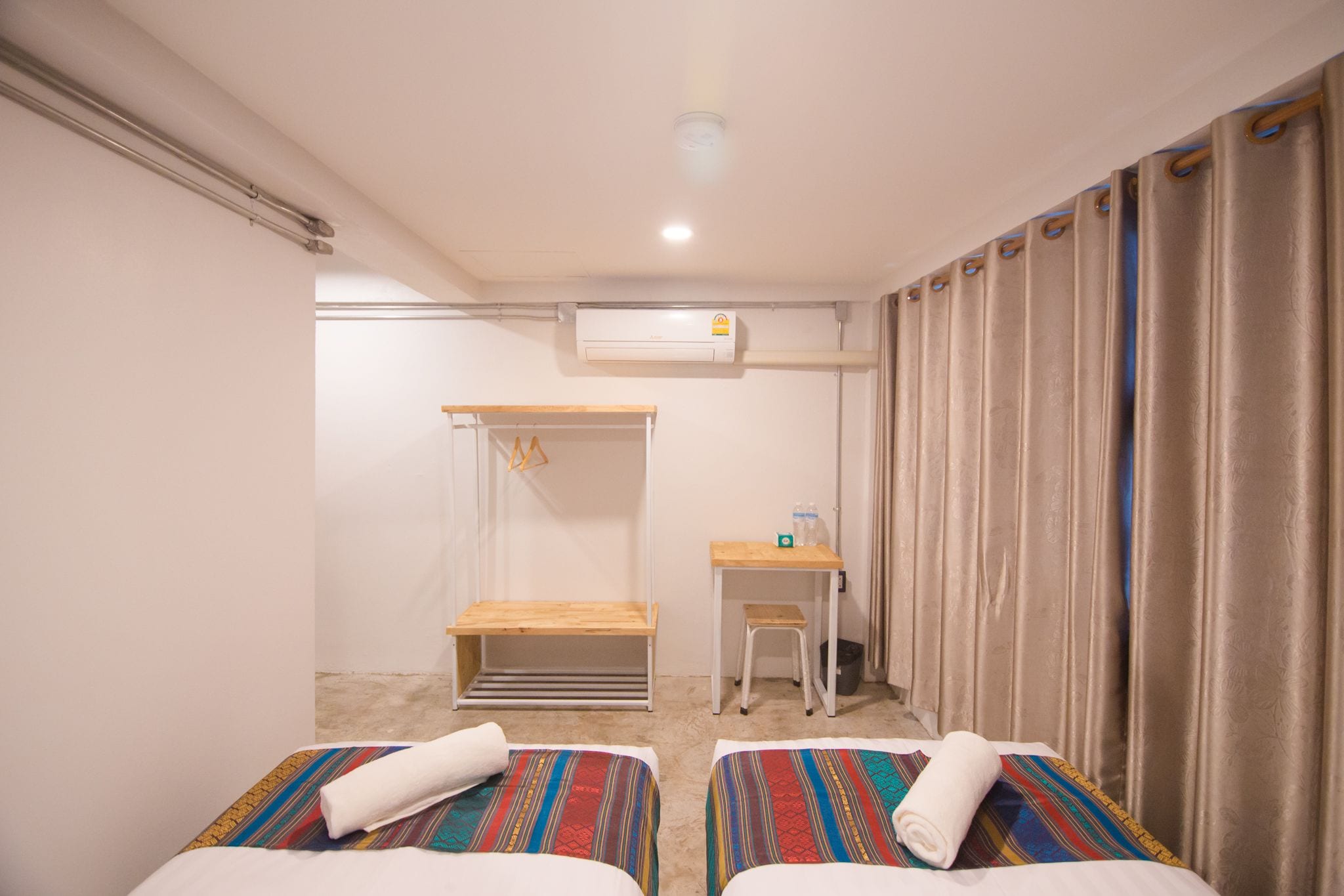 A hotel room with two beds, a small table, a bench, an air conditioning unit, and a curtain.