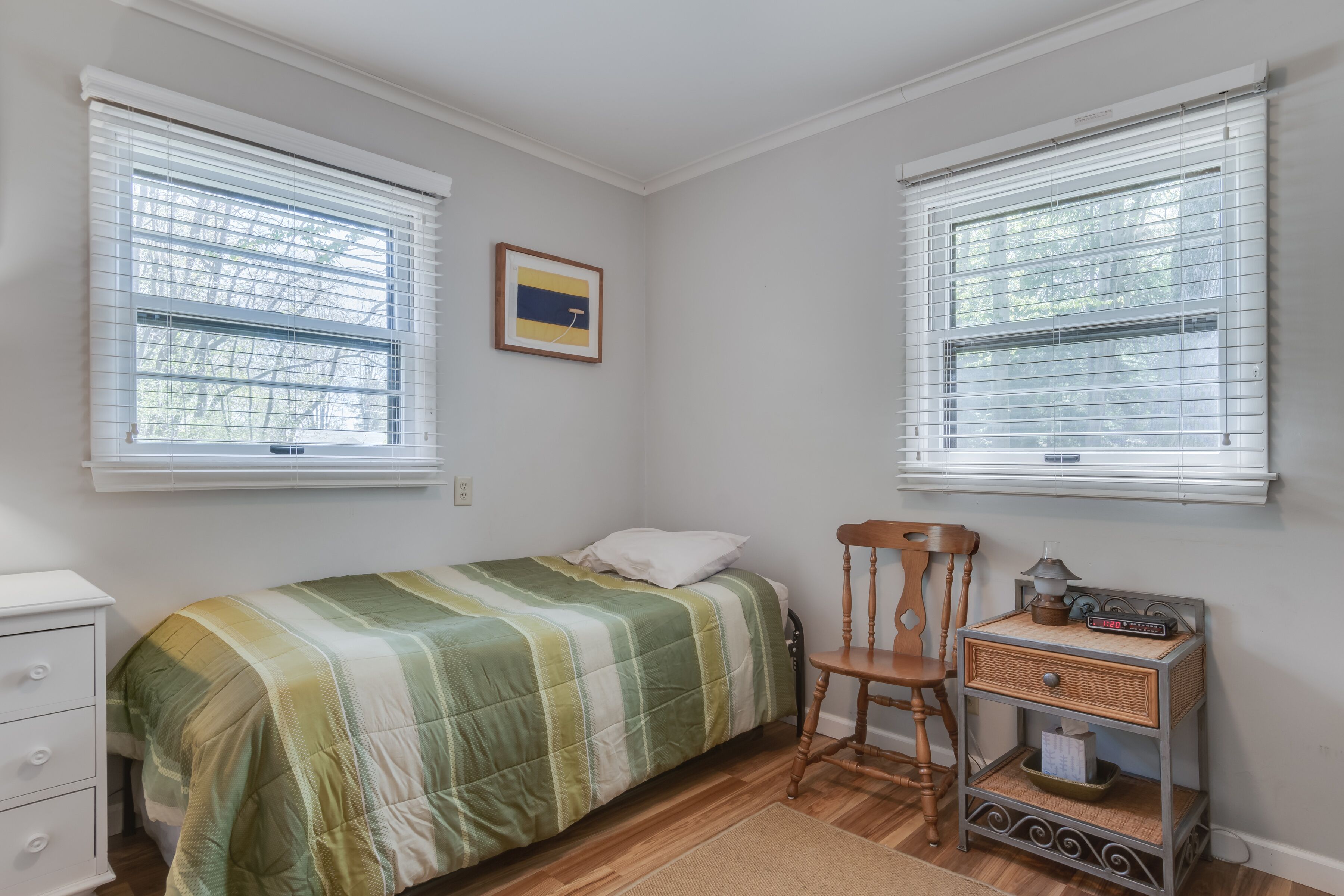 Une chambre à coucher comprenant un lit, une chaise, une table de chevet et deux fenêtres équipées de stores.