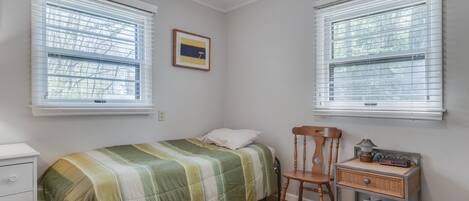 Une chambre à coucher comprenant un lit, une chaise, une table de chevet et deux fenêtres équipées de stores.