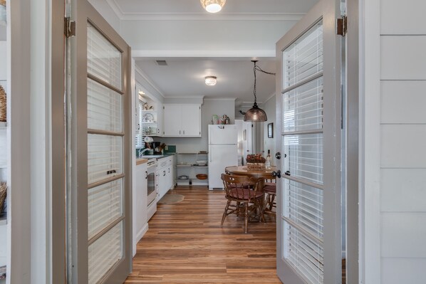 A narrow hallway with wooden flooring leading to a kitchen area with white cabinets, a refrigerator, and a dining table with chairs.