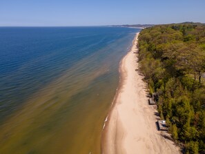 An aerial view of a beach with a clear sky, a sandy shore, and dense greenery on the right side.