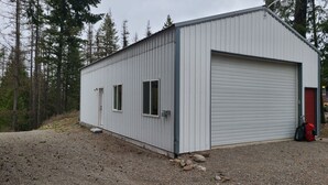 A single-story metal building with a garage door, two windows, and a red door. - Included RV Spot + 10 min to Silverwood  (Hayden)