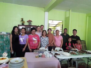 A group of people gathered around a table with various dishes, in a room with green walls and a window.