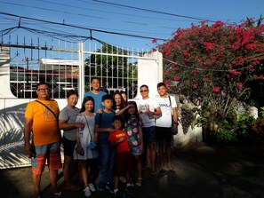 A group of people standing in front of a white gate with decorative metal bars, with a lush garden and flowering plants in the background.
