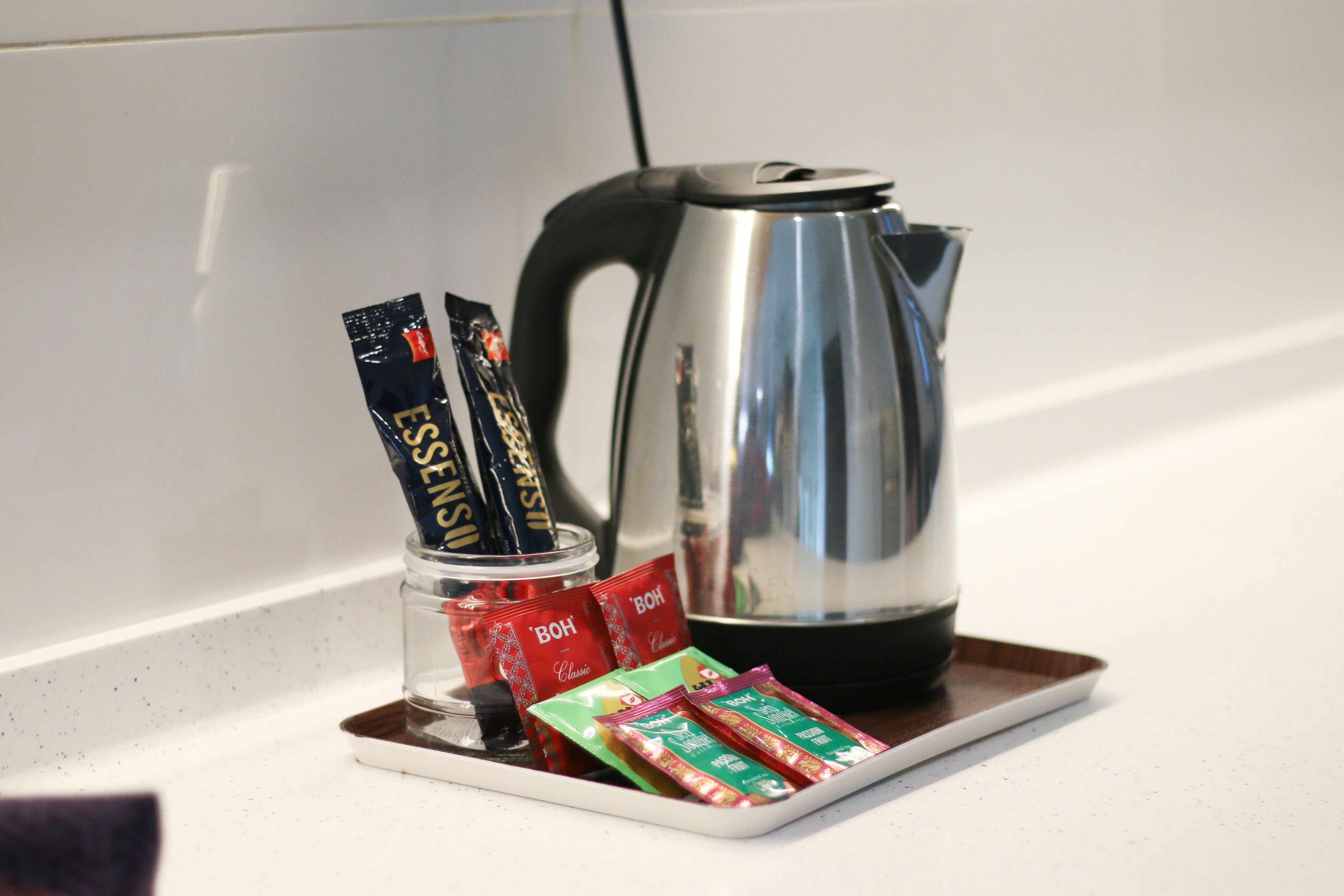 Un plateau sur lequel se trouvent une bouilloire électrique argentée, ainsi que des sachets de café et de thé, le tout posé sur un comptoir blanc.