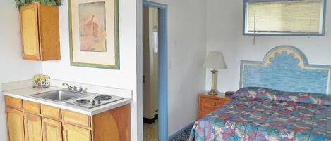 Quarto de hotel compacto com cozinha compacta, uma cama com colcha colorida, mesa de cabeceira com abajur e um quadro na parede.