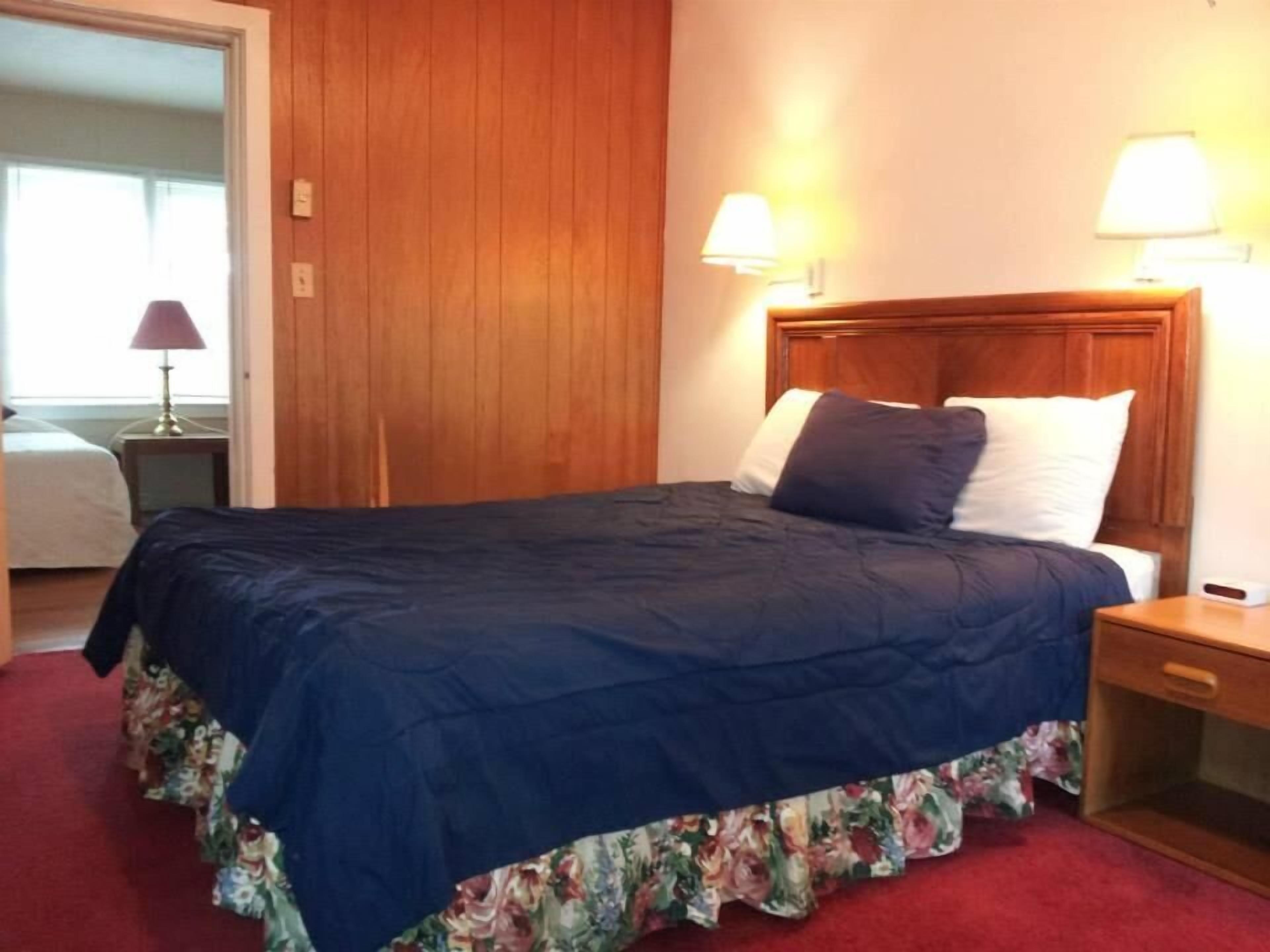 A neatly made bed with a blue bedspread and floral skirt, two bedside lamps, and a wooden headboard in a hotel room.