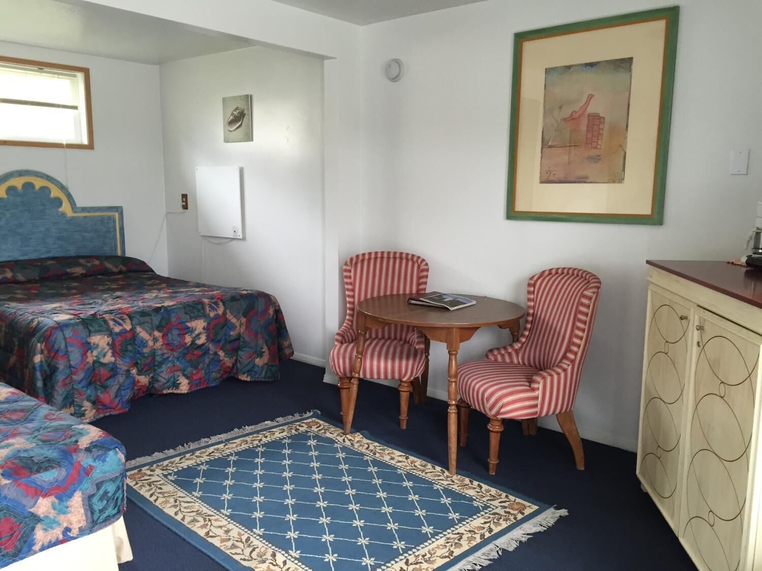 A room with a bed, two striped chairs, a small table, a cabinet, and a rug.