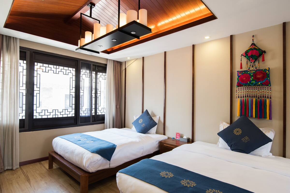 Une chambre d’hôtel avec deux lits, un plafond en bois, une fenêtre ornée d’un motif géométrique et un élément décoratif suspendu agrémenté de glands.