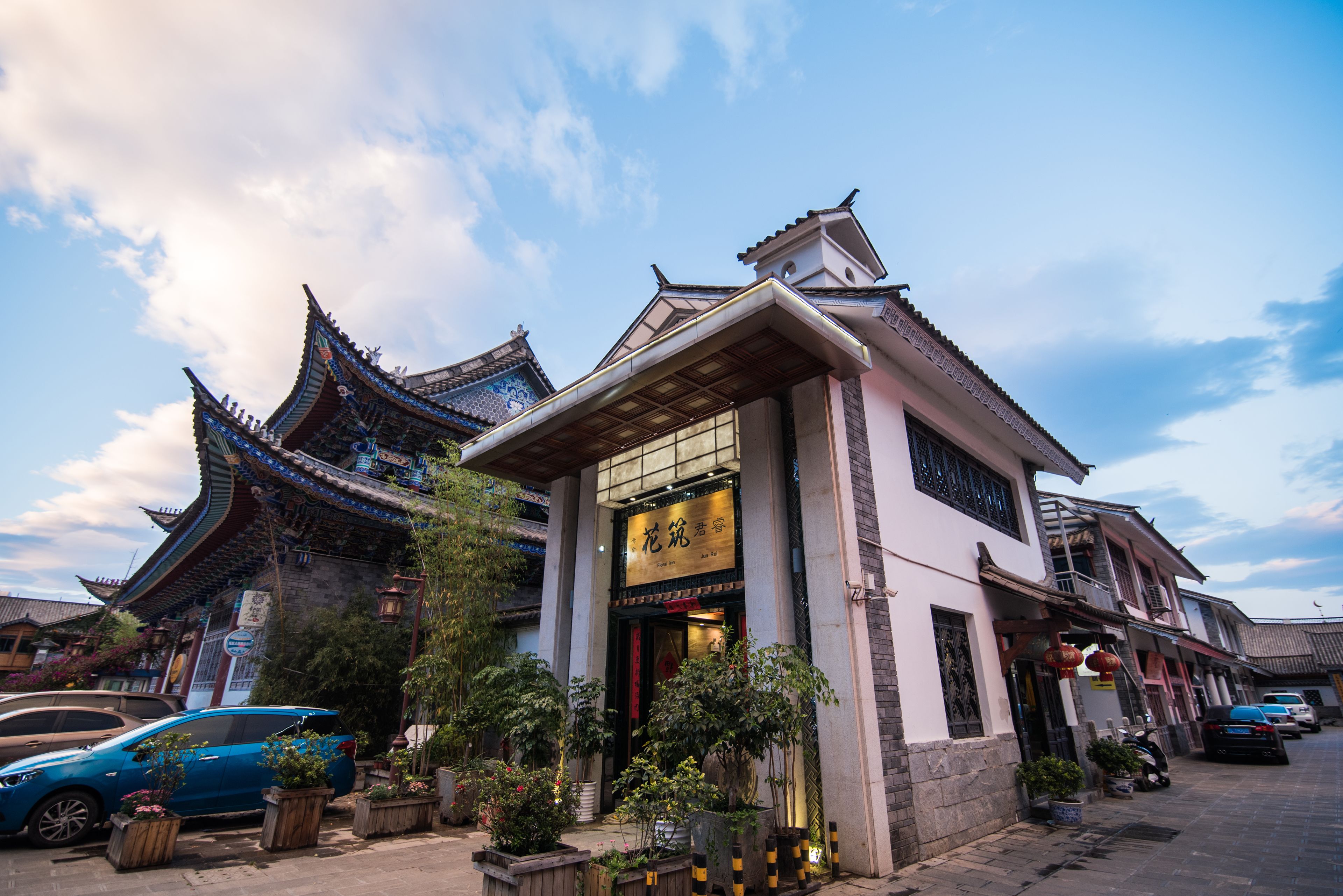 A traditional building with a sign in a foreign language, surrounded by potted plants and parked cars.