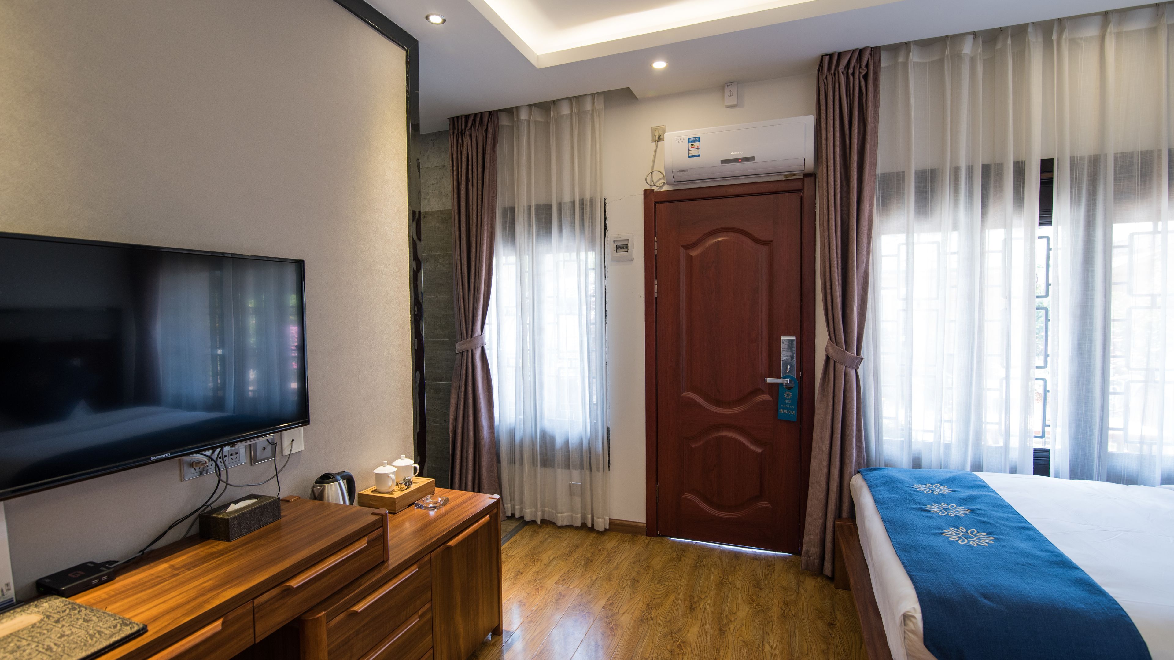 A hotel room with a flat-screen TV, wooden furniture, a bed with a blue blanket, and a door with a keycard lock.