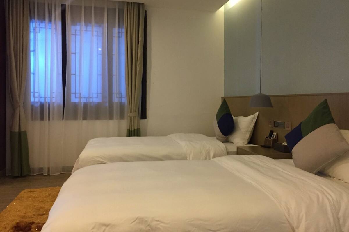 A hotel room with two beds, a nightstand, a lamp, and a window with curtains.