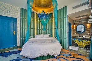 Une chambre à coucher avec un lit, un lustre, une salle de bain avec un lavabo et un miroir, et un tapis à motifs.
