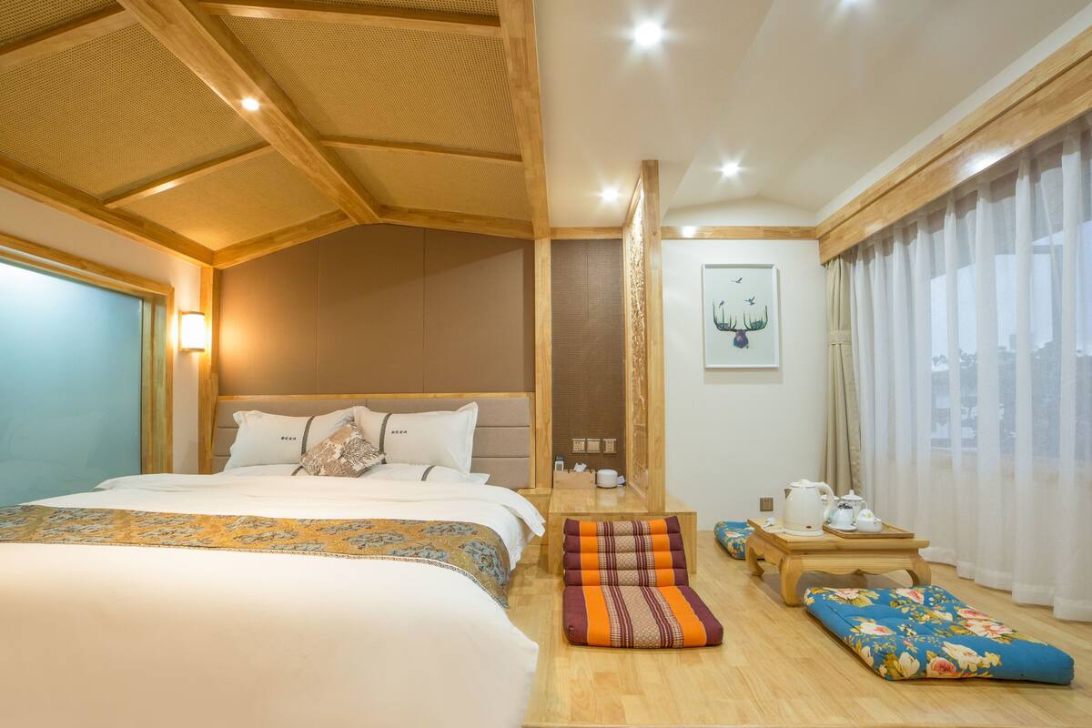 A hotel room with a large bed, a wooden ceiling, a small table with tea set, and a window with sheer curtains.