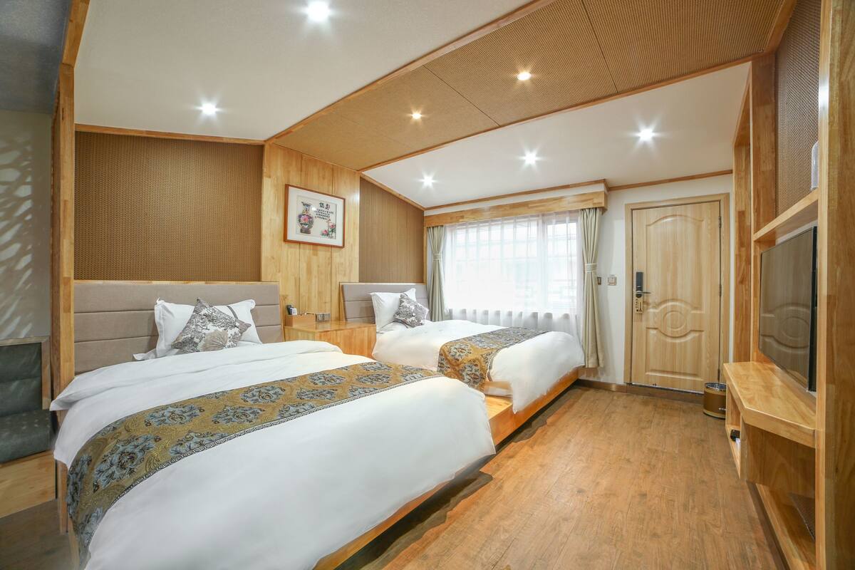 A hotel room with two beds, wooden flooring, a television, and a large window with curtains.