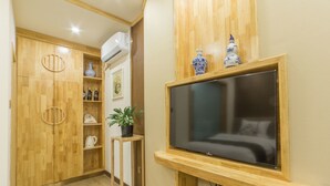 Habitación de hotel con un armario de madera, un televisor montado en la pared, una pequeña mesa con una planta y un estante con dispositivos electrónicos.