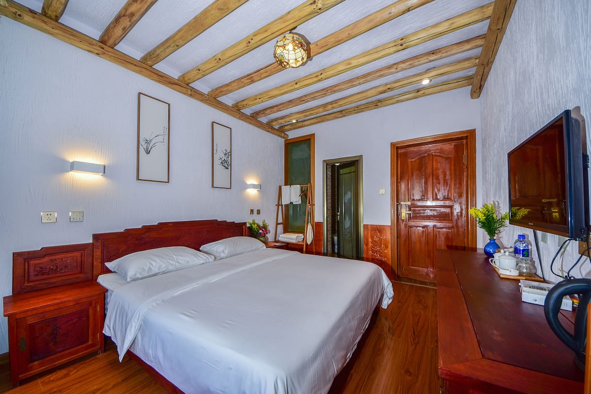 Een slaapkamer met een houten plafond, een bed met witte lakens, een houten nachtkastje, een bureau met een stoel en een spiegel.
