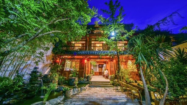 A traditional building with a tiled roof, a stone staircase leading to the entrance, and a well-maintained garden with various plants and a small pond.