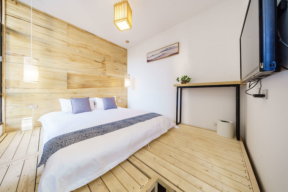 Ein modernes Schlafzimmer mit Holzboden, einem großen Bett mit weißer und blauer Bettwäsche, einem Holz-Kopfende, einem an der Wand befestigten Fernseher und einer kleinen Pflanze auf einem Regal.