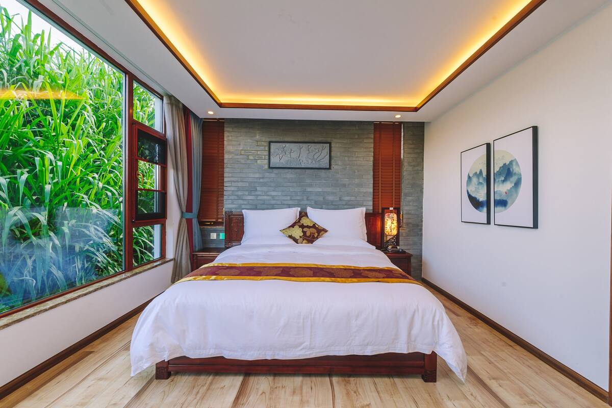 Ein geräumiges Schlafzimmer mit einem großen Bett, Blick auf Grün durch ein Fenster, einer Wand mit Steinverkleidung und Kunstwerken an den Wänden.