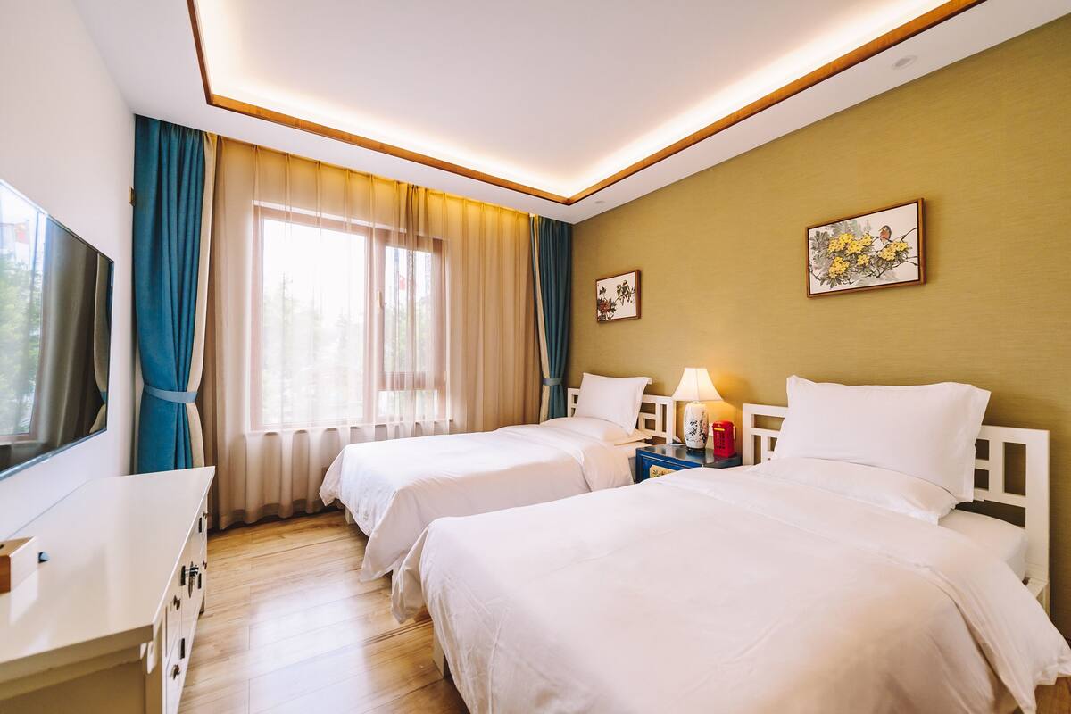 Ein Hotelzimmer mit zwei Betten, einem Fernseher, einem Nachttisch und einem Fenster mit Vorhängen.