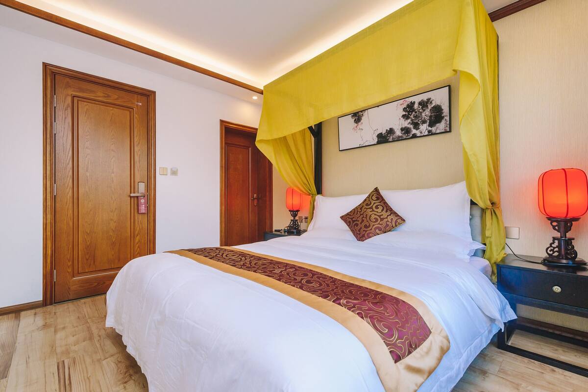 Ein Hotelzimmer mit einem großen Bett, einer Holztür, einem Nachttisch mit einer roten Lampe und einem gelben Baldachin über dem Bett.