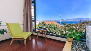 Ein Zimmer mit Holzboden, einem grünen Sessel, einem kleinen Holztisch mit einer Flasche und Gläsern sowie Blick auf das Meer und die Berge.