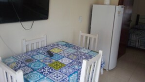 A dining area with a table covered in a patterned tablecloth, white chairs, a white refrigerator, and a television mounted on the wall.