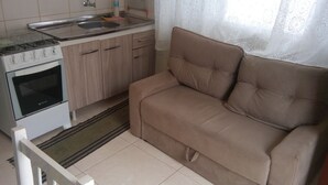 A compact kitchen with a stove, oven, and sink. A beige sofa is positioned near a window with lace curtains.