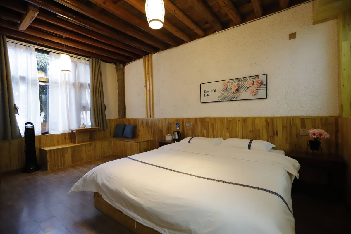 A bedroom with a large bed, wooden paneling, a window with curtains, a bedside table, and a wall-mounted artwork.