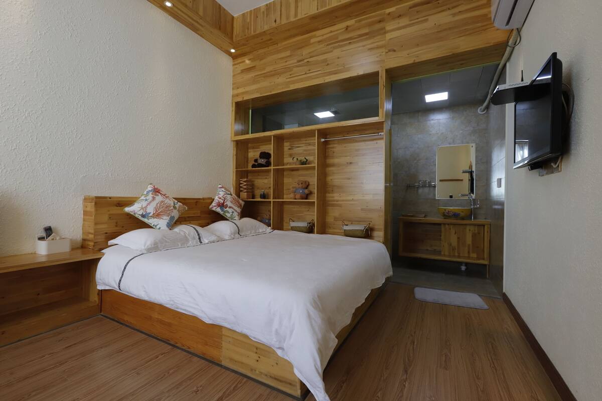 A bedroom with a wooden headboard, a bed with white bedding, a wooden shelf unit with decorative items, a television mounted on the wall, and a small bathroom with a mirror and sink.