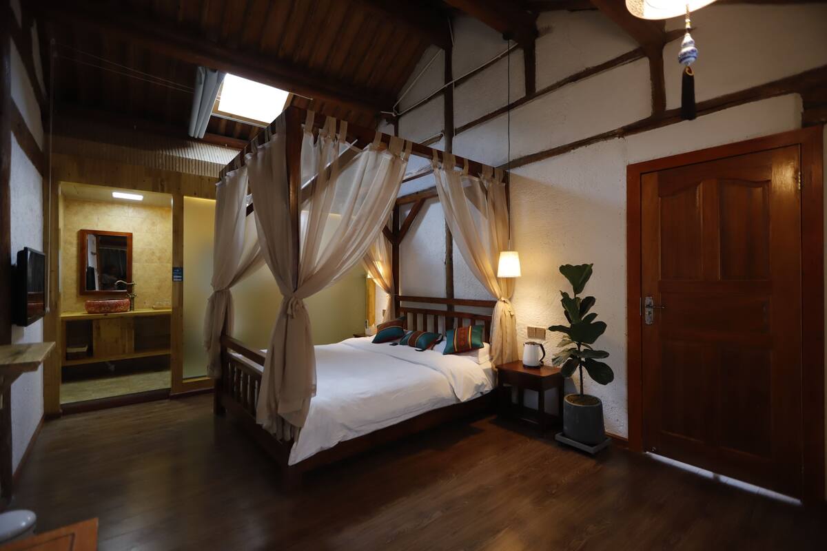 A bedroom with a four-poster bed, wooden flooring, a nightstand with a lamp, and a door.
