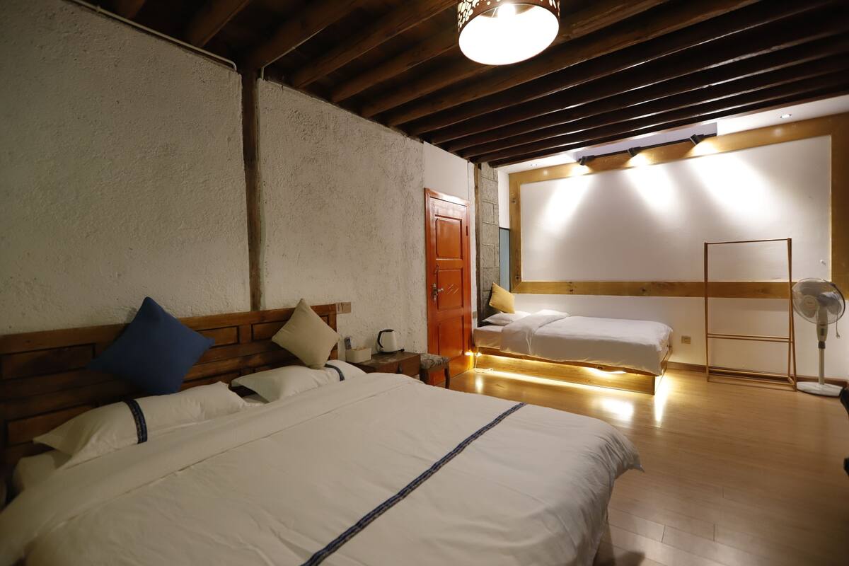 A hotel room with two beds, wooden headboards, a wooden floor, a ceiling fan, and a door leading to another room.