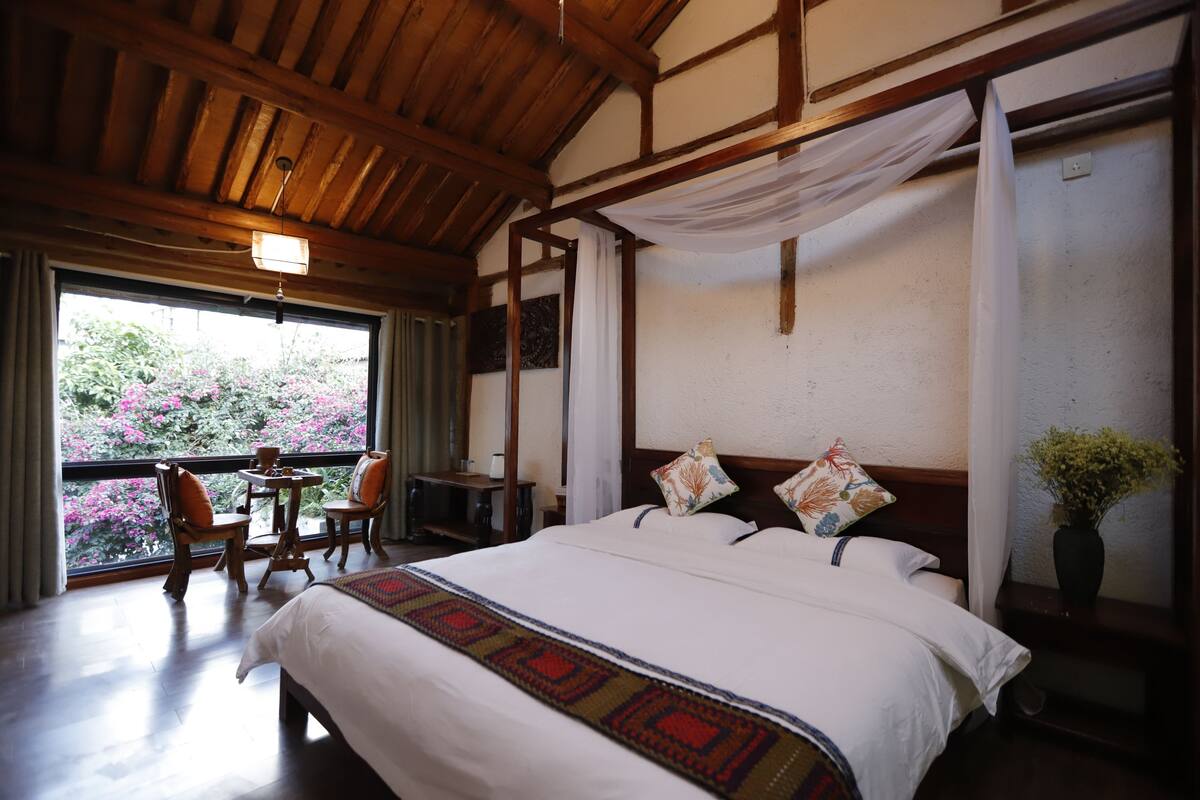 A bedroom with a large bed, wooden ceiling, a small table with chairs, and a window with a view of pink flowers.