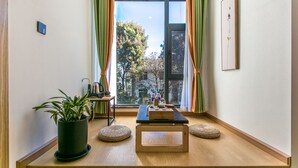 Ein kleines Zimmer mit Holzboden, einem Tisch mit Wasserkocher und Tassen, einer Topfpflanze und Blick auf Bäume durch ein Fenster mit Vorhängen.