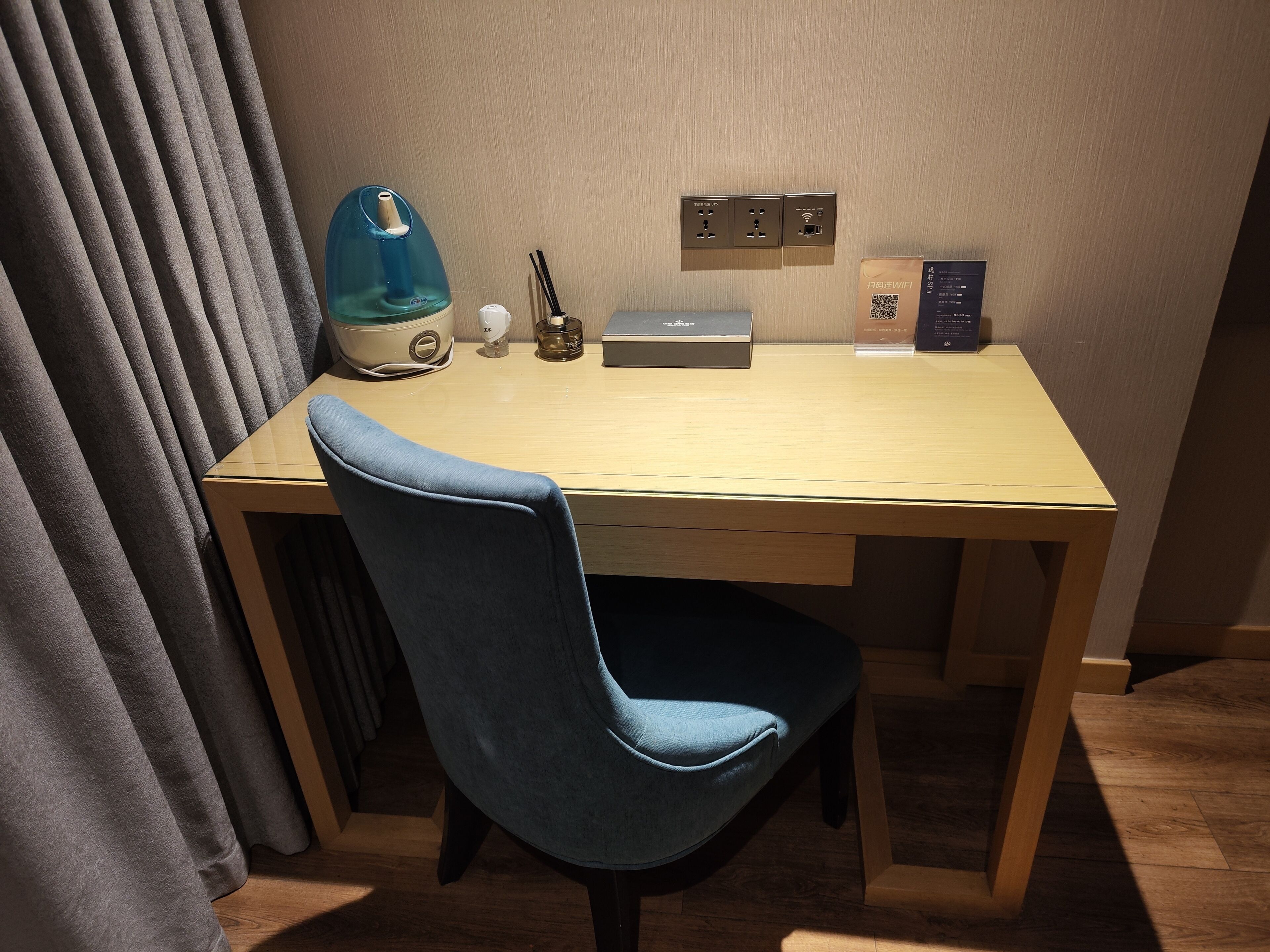 A hotel room with a wooden desk, a blue chair, a kettle, a diffuser, and a power outlet with a Wi-Fi symbol.