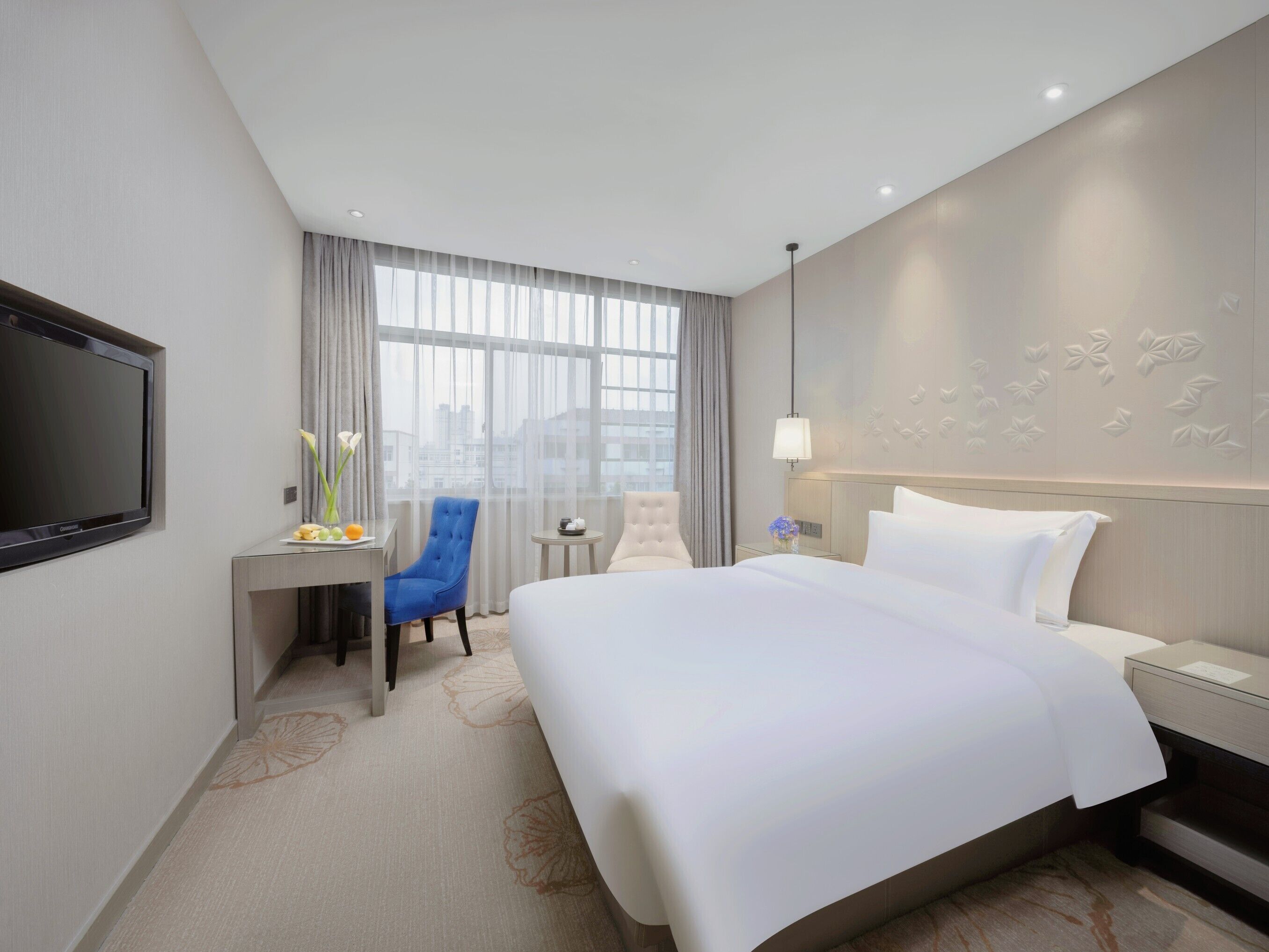 Habitación de hotel con una cama grande, un escritorio con una silla, un televisor y vistas a la ciudad a través de amplios ventanales.