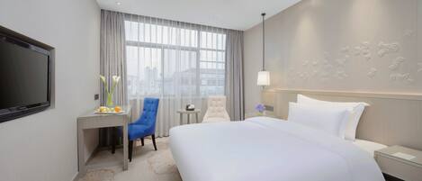 A hotel room with a large bed, a desk with a chair, a TV, and a view of the city through large windows.