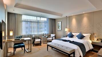 A hotel room with a large bed, a desk, two chairs, a small table, and a large window with curtains.