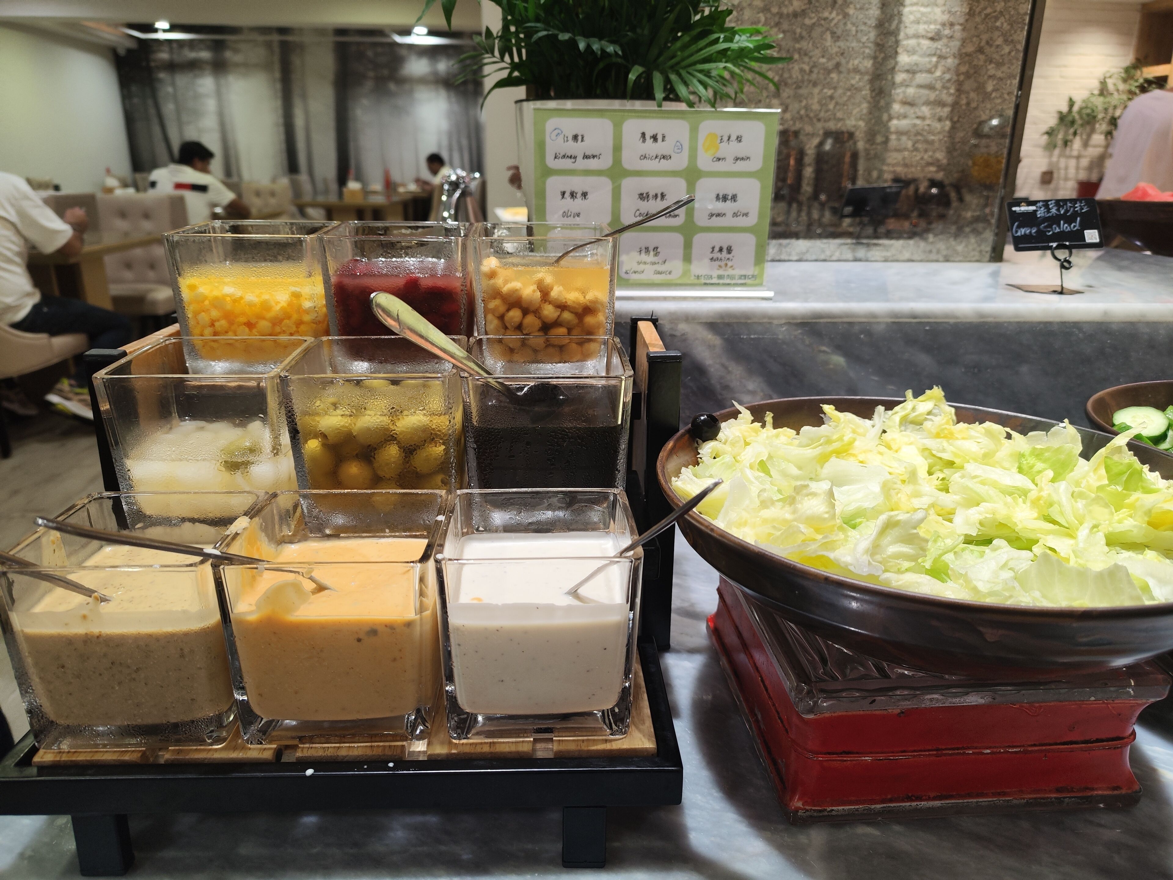Un bufé con diversas opciones de comida, incluyendo un tazón de ensalada de lechuga y varios recipientes con diferentes salsas y aderezos.