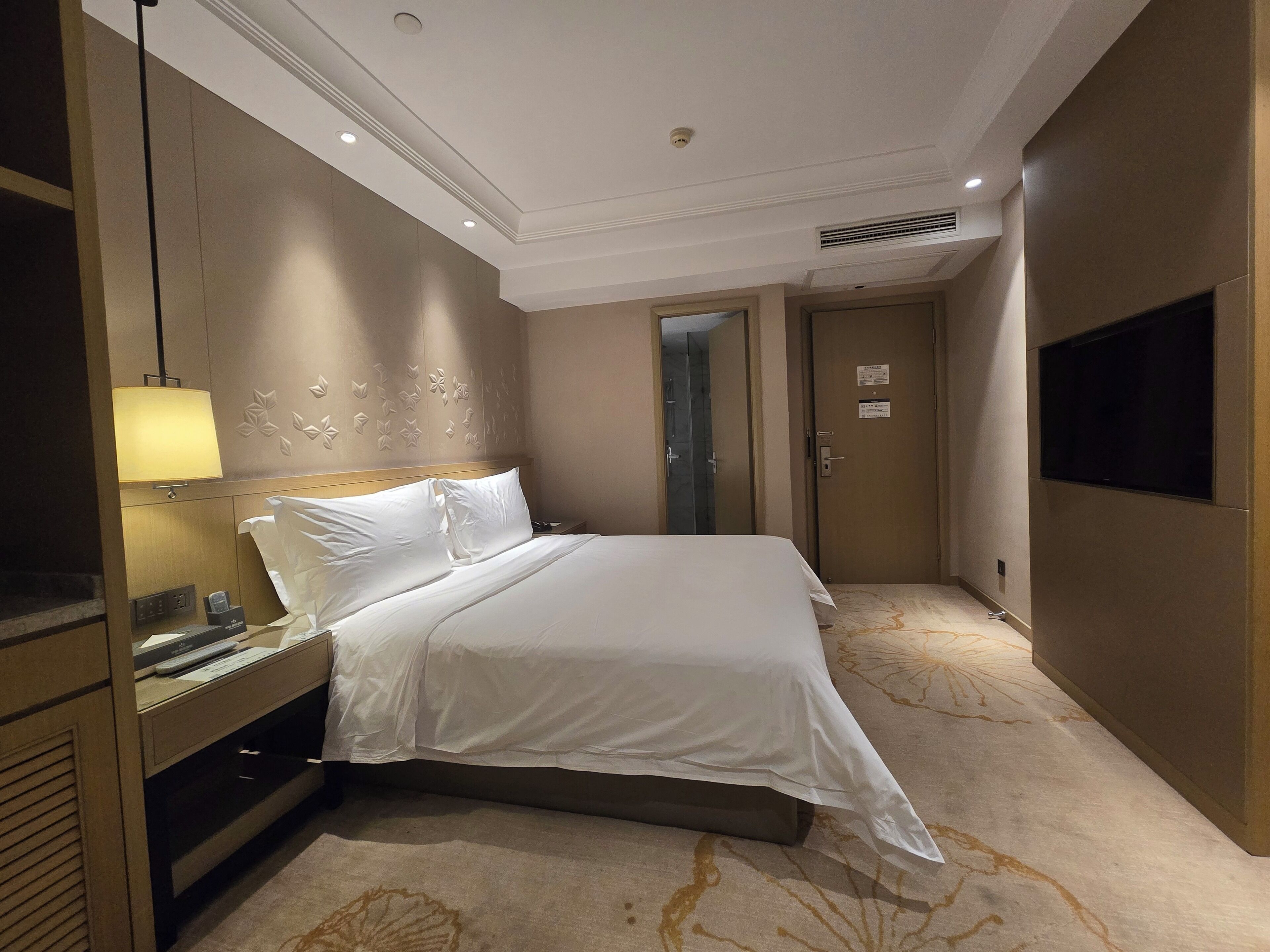 A hotel room with a bed, bedside table, lamp, and a television on the wall.