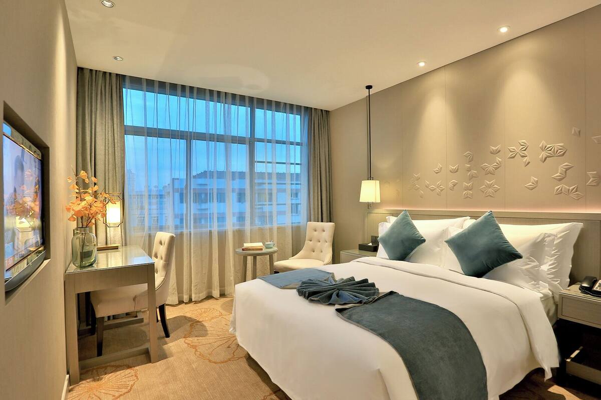 Een hotelkamer met een groot bed, een bureau, een stoel, een nachtkastje en uitzicht op de stad door grote ramen.